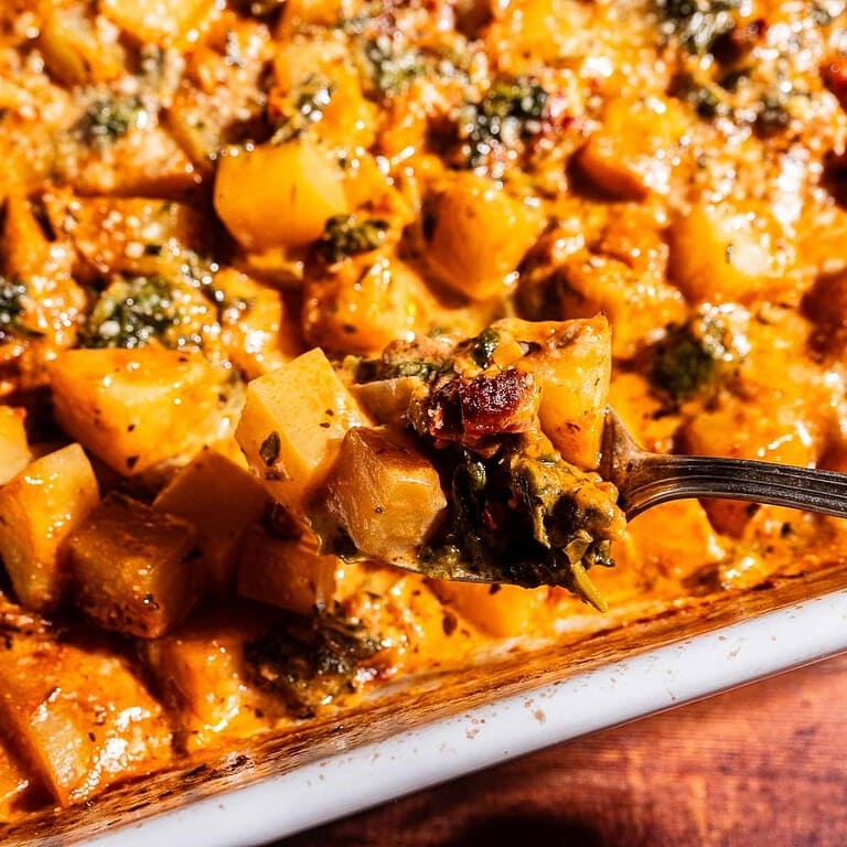 A close-up of a baked casserole featuring cubed vegetables, greens, and a creamy, browned sauce reminiscent of marry me potatoes. A fork lifts a bite-sized portion above the dish.