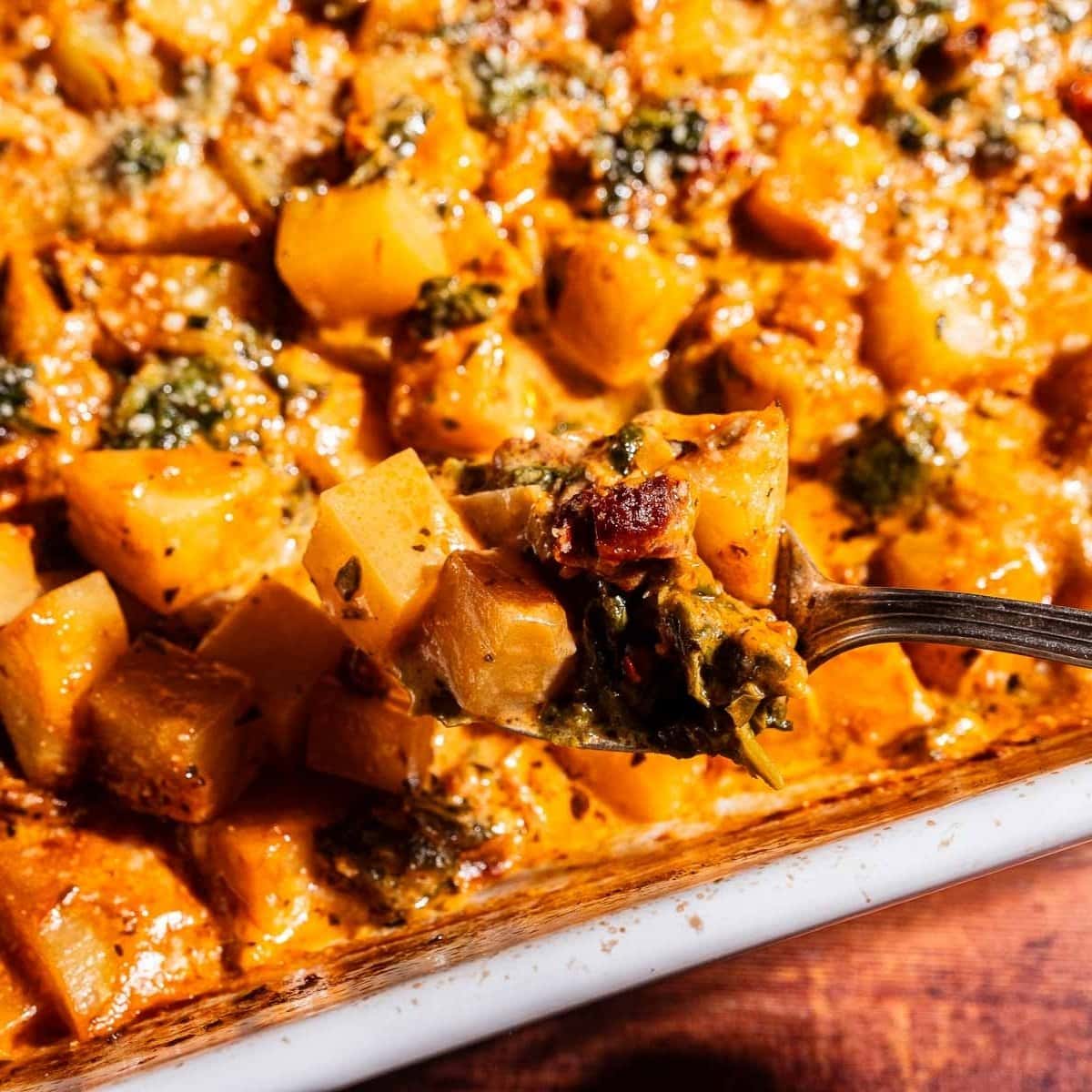 A close-up of a baked casserole featuring cubed vegetables, greens, and a creamy, browned sauce reminiscent of marry me potatoes. A fork lifts a bite-sized portion above the dish.
