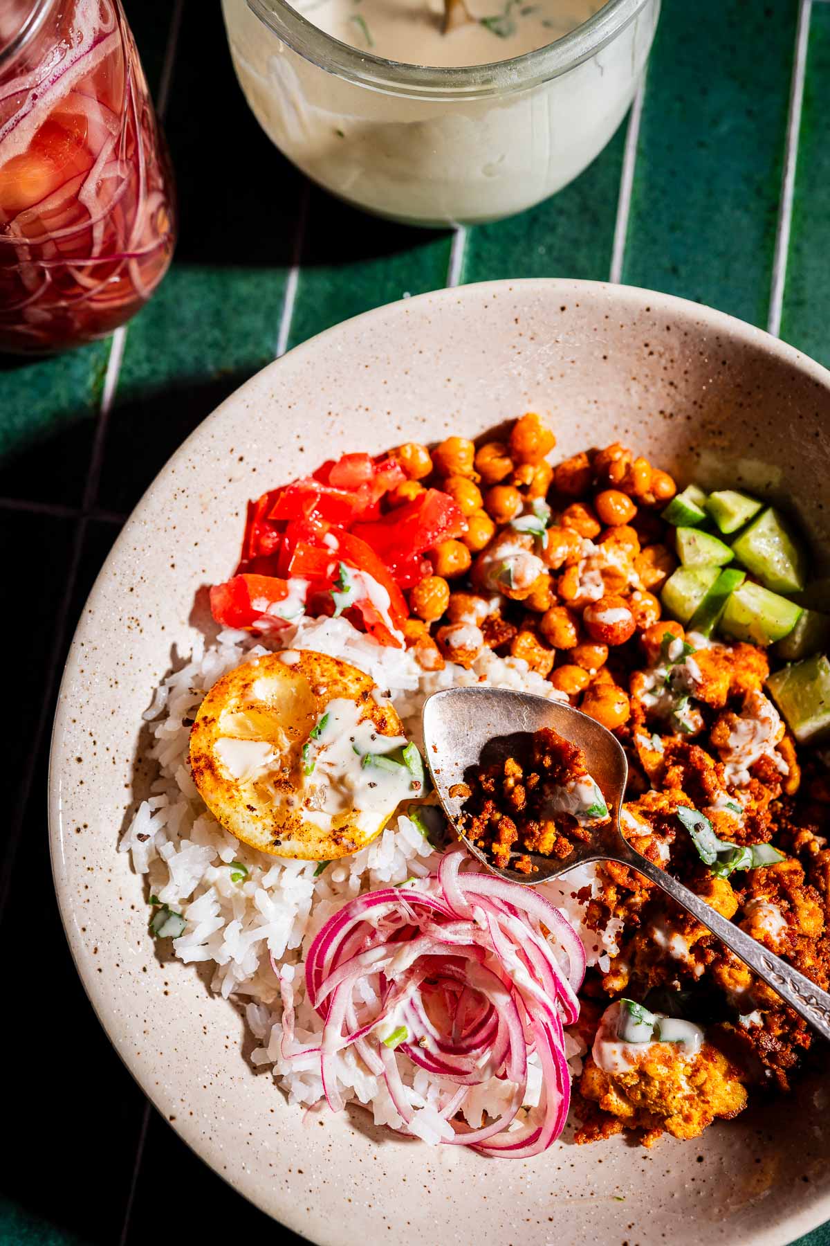 A bowl with rice, tofu shawarma, roasted chickpeas, sliced cucumbers, tomatoes, red onions, a roasted lemon slice, sauce, and a jar of dressing in the background.