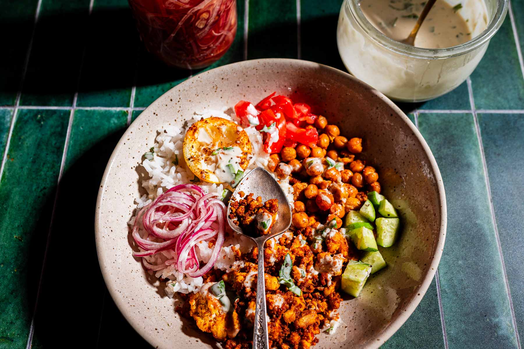 A bowl with rice, spiced chickpeas, tofu shawarma, diced cucumber, tomato, pickled onions, a slice of lemon, and creamy sauce, with a spoon placed in the center.