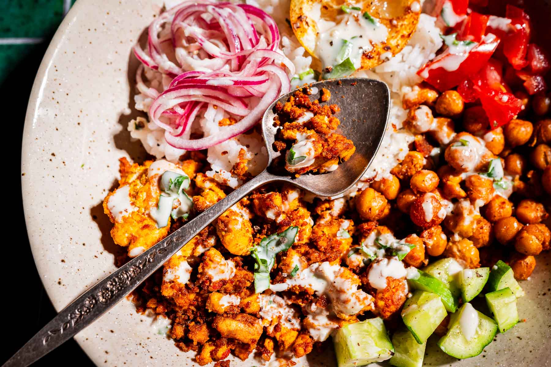 A plate with spiced chickpeas, diced cucumber, sliced red onions, tomatoes, rice, and tofu shawarma, finished with a creamy white sauce and a lemon wedge, with a spoon resting on top.