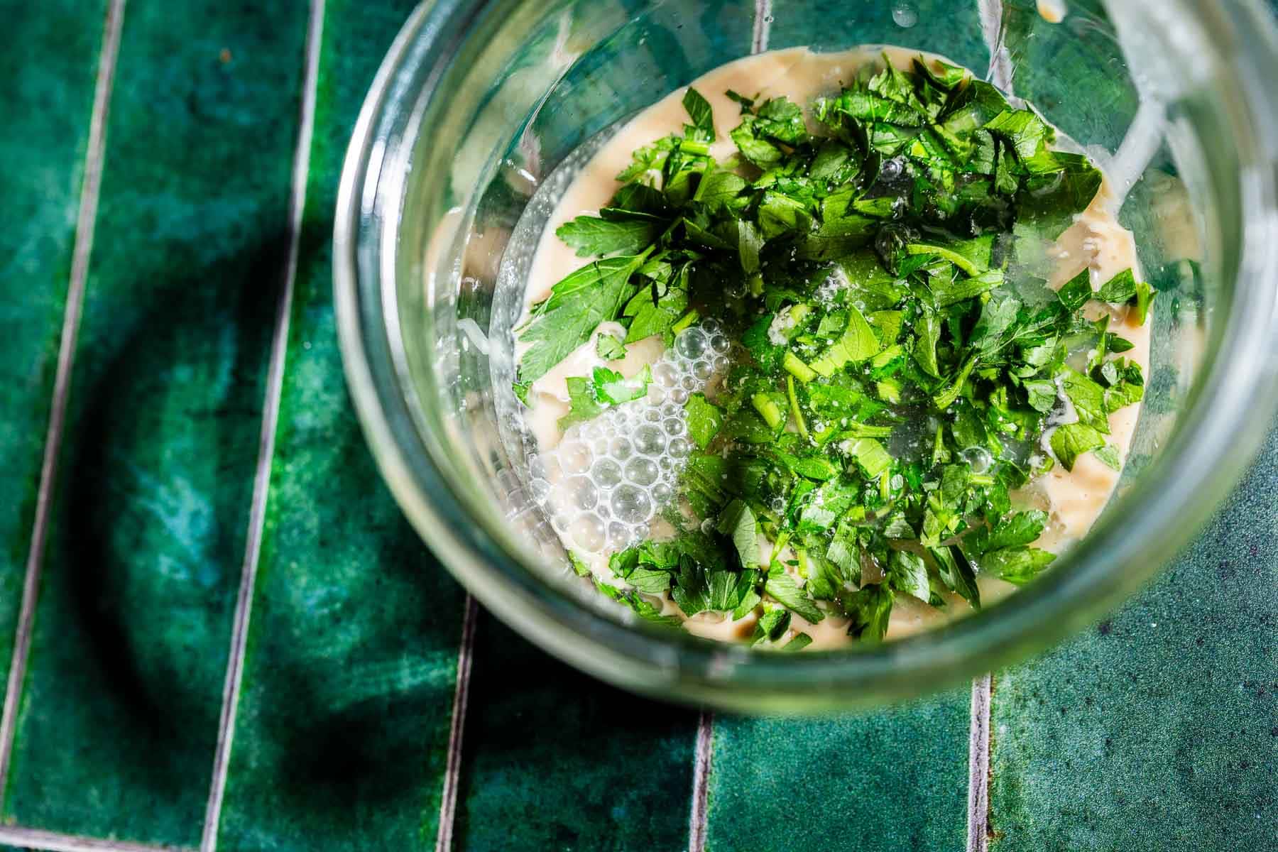 A glass bowl containing chopped parsley, yeast, and liquid ingredients for tofu shawarma sits on a green tiled surface.