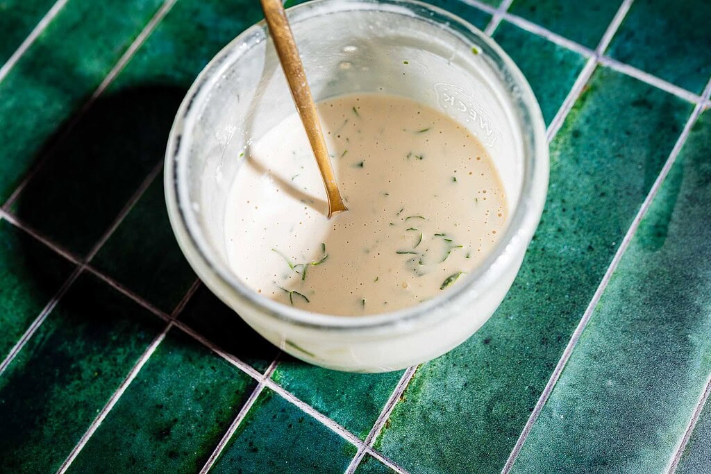 A glass bowl of creamy dressing with herbs, perfect for drizzling over tofu shawarma, is mixed with a wooden spoon and sits on a green tiled surface.