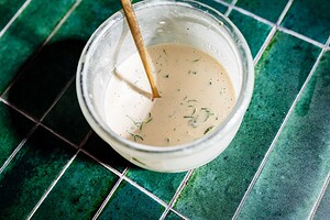 A glass bowl of creamy dressing with herbs, perfect for drizzling over tofu shawarma, is mixed with a wooden spoon and sits on a green tiled surface.