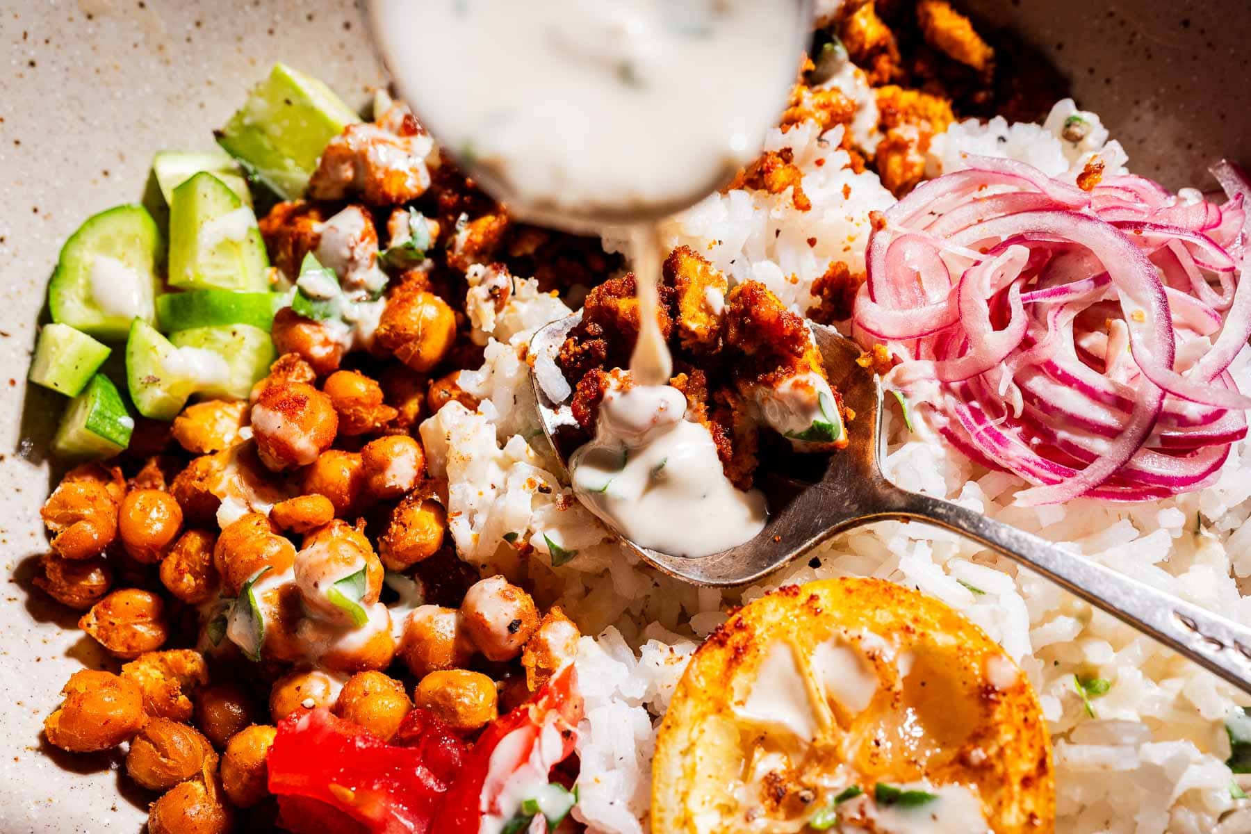 A close-up of a bowl with rice, chickpeas, cucumber, tomato, sliced red onion, roasted lemon, and tofu shawarma, as a spoon drizzles sauce on top.