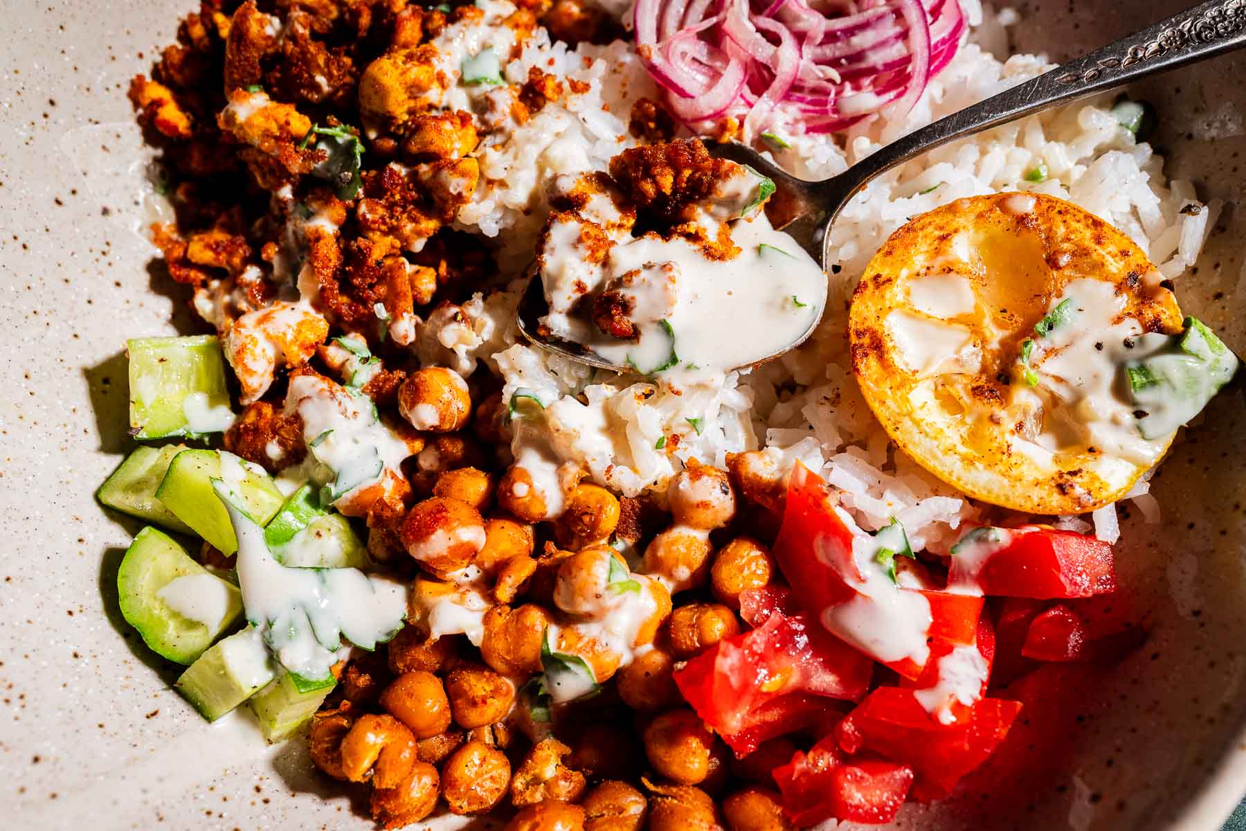 A bowl with rice, roasted chickpeas, chopped cucumber, tomatoes, sliced red onions, lemon, tofu shawarma crumbles, and creamy sauce.