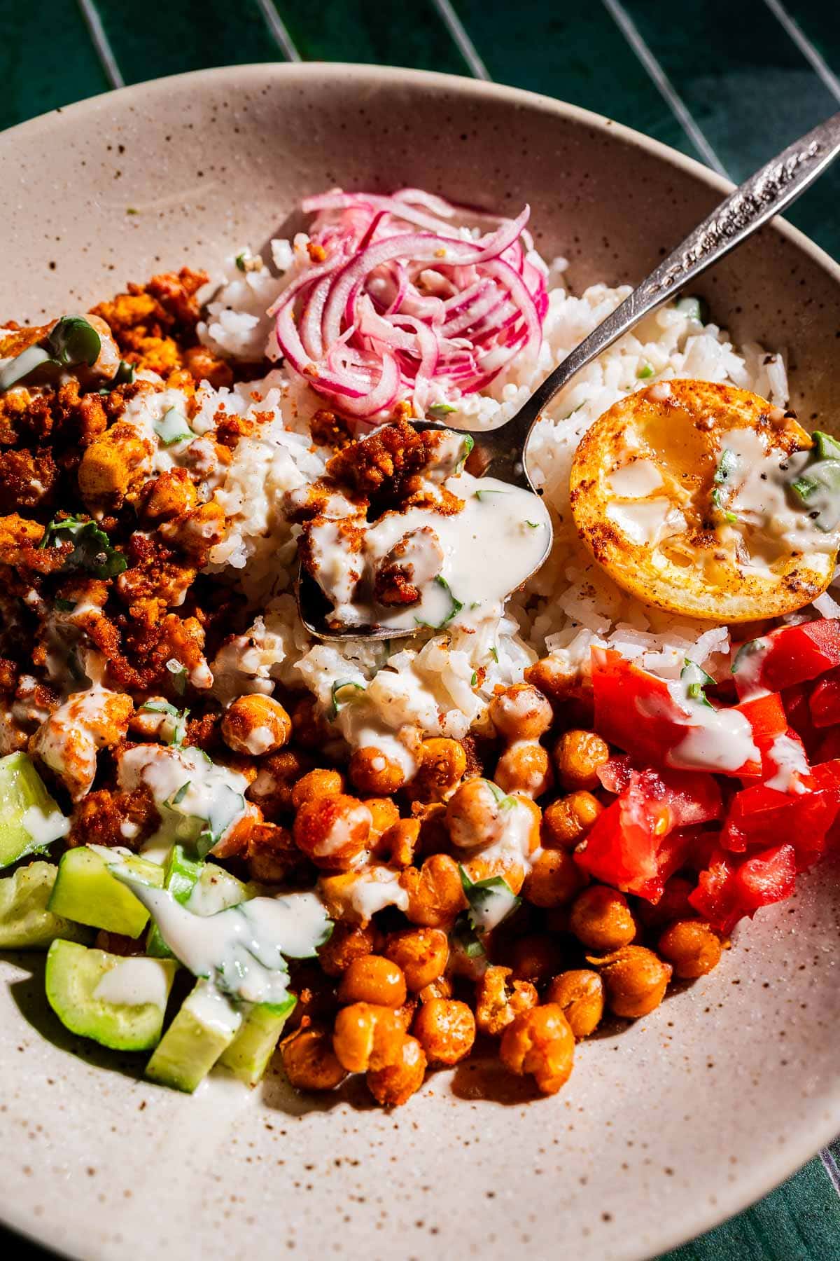A bowl of rice topped with chickpeas, chopped tomatoes, cucumbers, pickled onions, crumbled tofu shawarma, a grilled lemon slice, and drizzled with a white sauce.