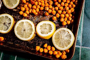 Sliced lemons, roasted chickpeas, and tofu shawarma on a dark, well-used baking tray resting on a green tiled surface.
