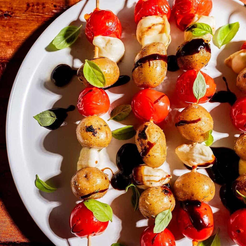 Skewers with roasted baby potatoes, cherry tomatoes, mozzarella, fresh basil, and balsamic glaze arranged on a white plate.