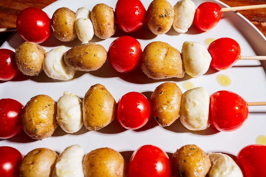 Wooden skewers with baby potatoes, cherry tomatoes, and mozzarella balls arranged on a white plate, drizzled with olive oil.