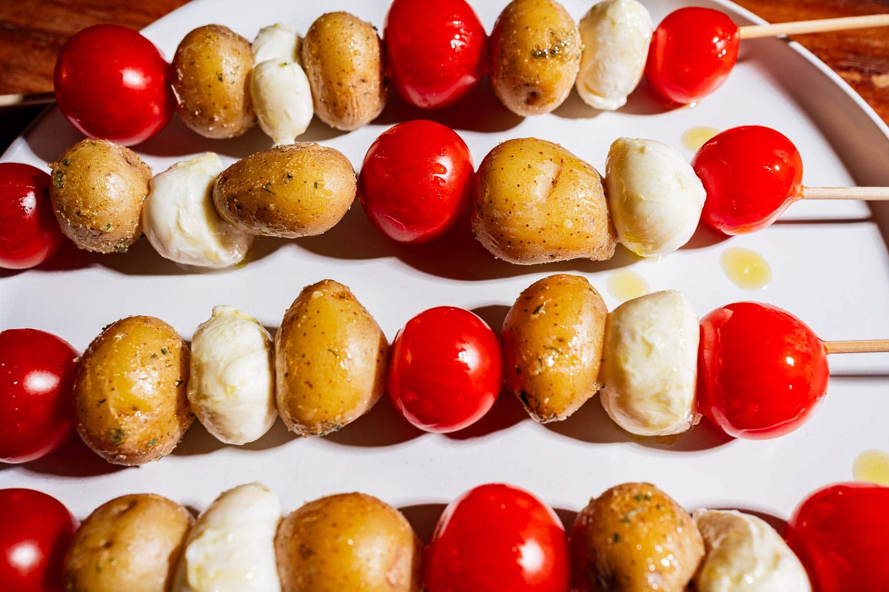 Wooden skewers with baby potatoes, cherry tomatoes, and mozzarella balls arranged on a white plate, drizzled with olive oil.