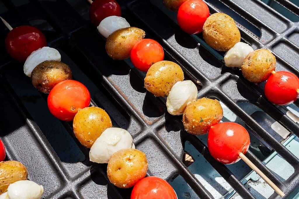 Skewers with cherry tomatoes, small potatoes, and mozzarella balls are being grilled on a barbecue.