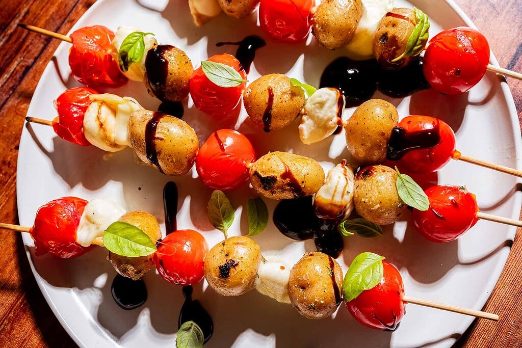 Skewers with cherry tomatoes, baby potatoes, and mozzarella balls, garnished with basil leaves and drizzled with balsamic glaze, served on a white plate.