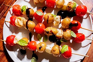Skewers with cherry tomatoes, baby potatoes, and mozzarella balls, garnished with basil leaves and drizzled with balsamic glaze, served on a white plate.