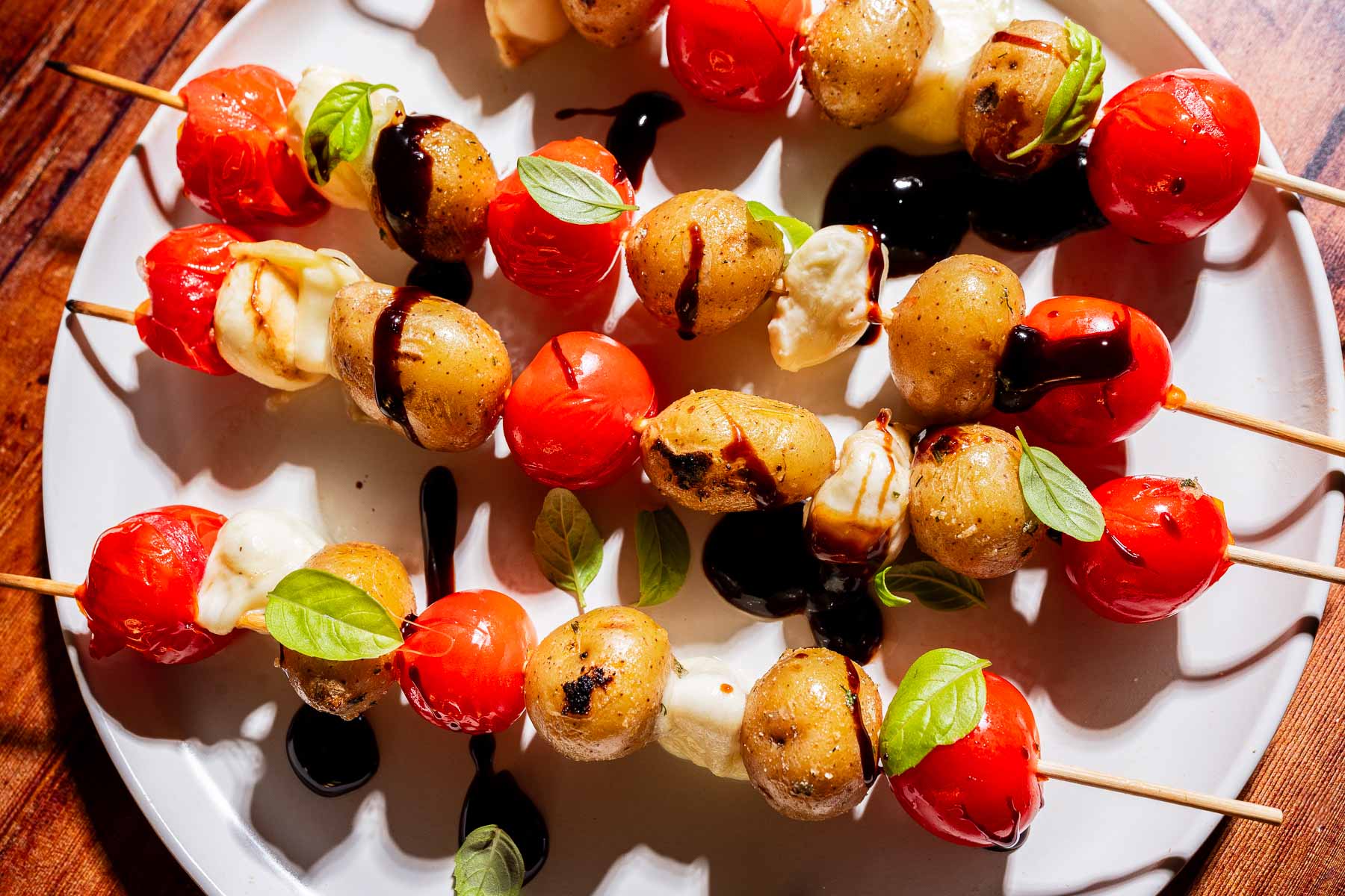 Skewers with cherry tomatoes, baby potatoes, and mozzarella balls, garnished with basil leaves and drizzled with balsamic glaze, served on a white plate.
