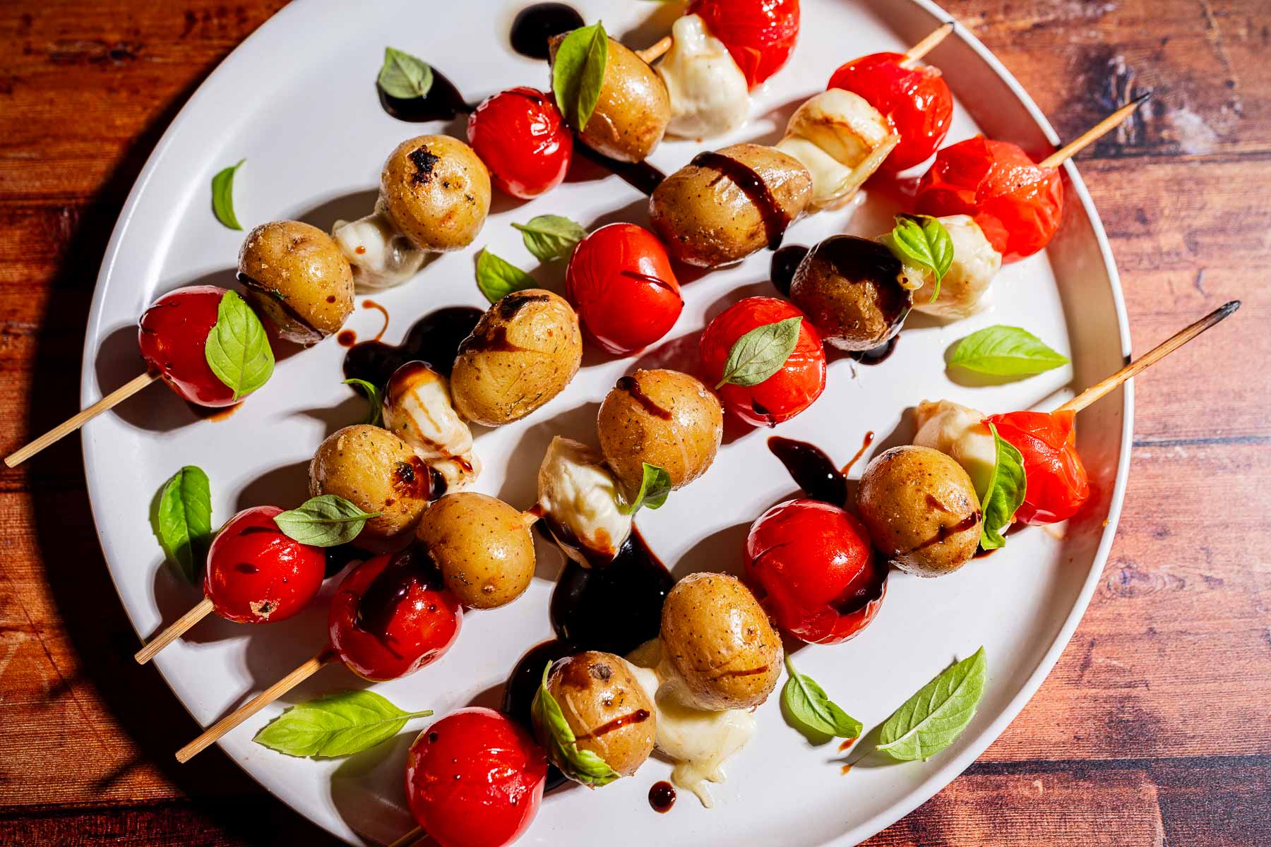 Skewers with cherry tomatoes, baby potatoes, mozzarella, and basil leaves drizzled with balsamic glaze on a white plate.