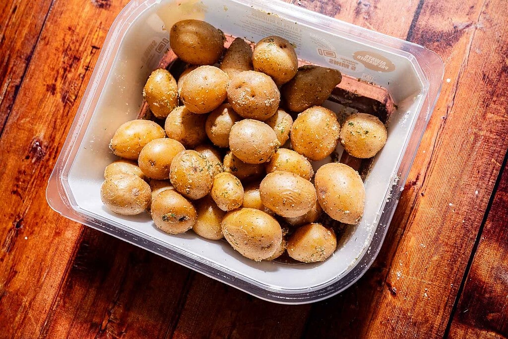 A plastic container filled with seasoned baby potatoes sits on a wooden surface.