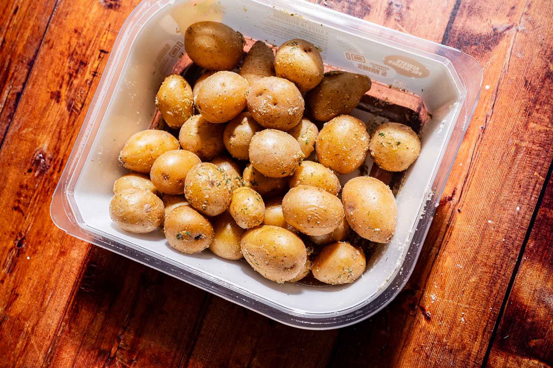 A plastic container filled with seasoned baby potatoes sits on a wooden surface.
