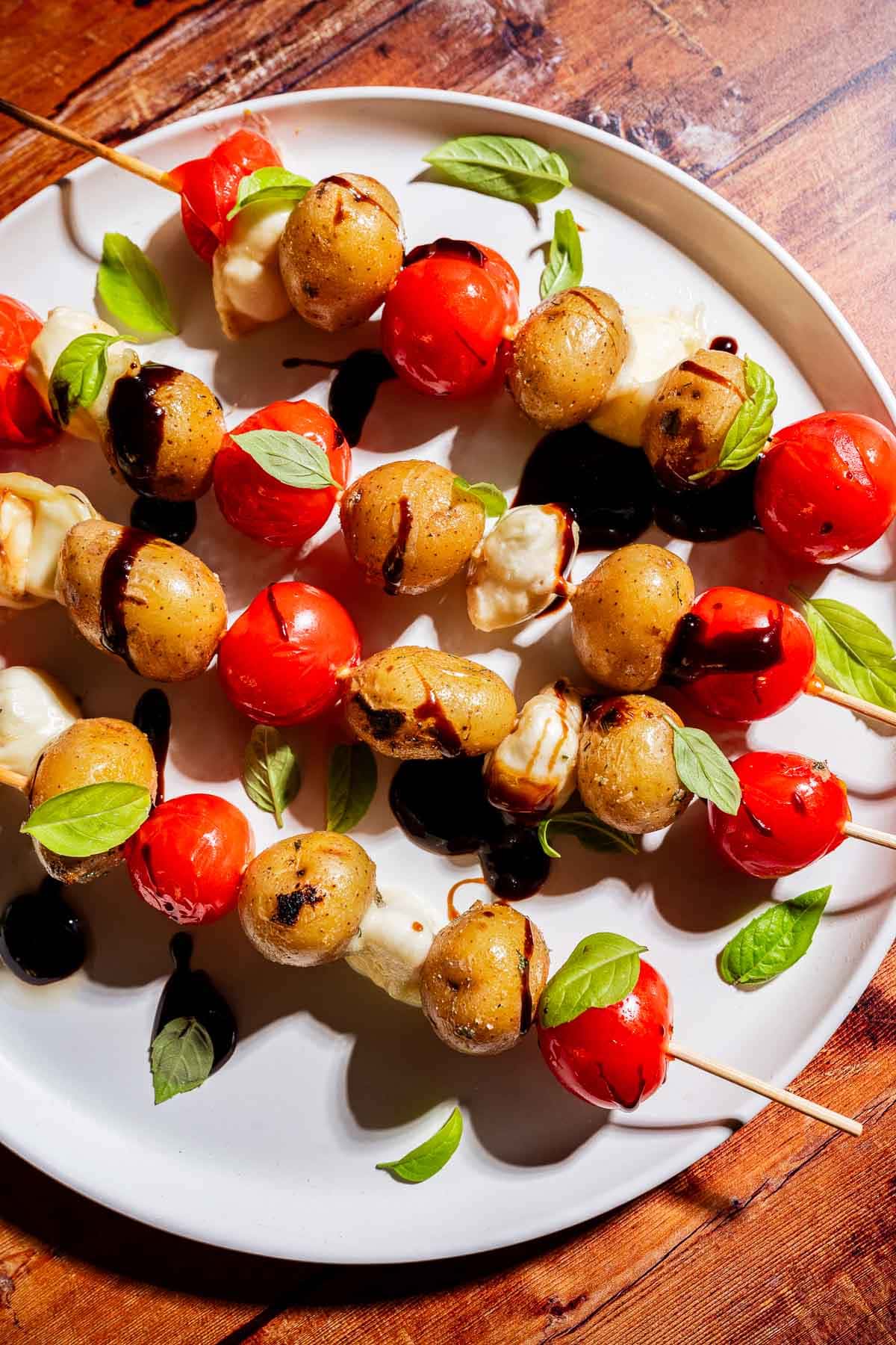 Skewers with cherry tomatoes, mozzarella balls, and small potatoes, garnished with basil leaves and drizzled with balsamic glaze on a white plate.