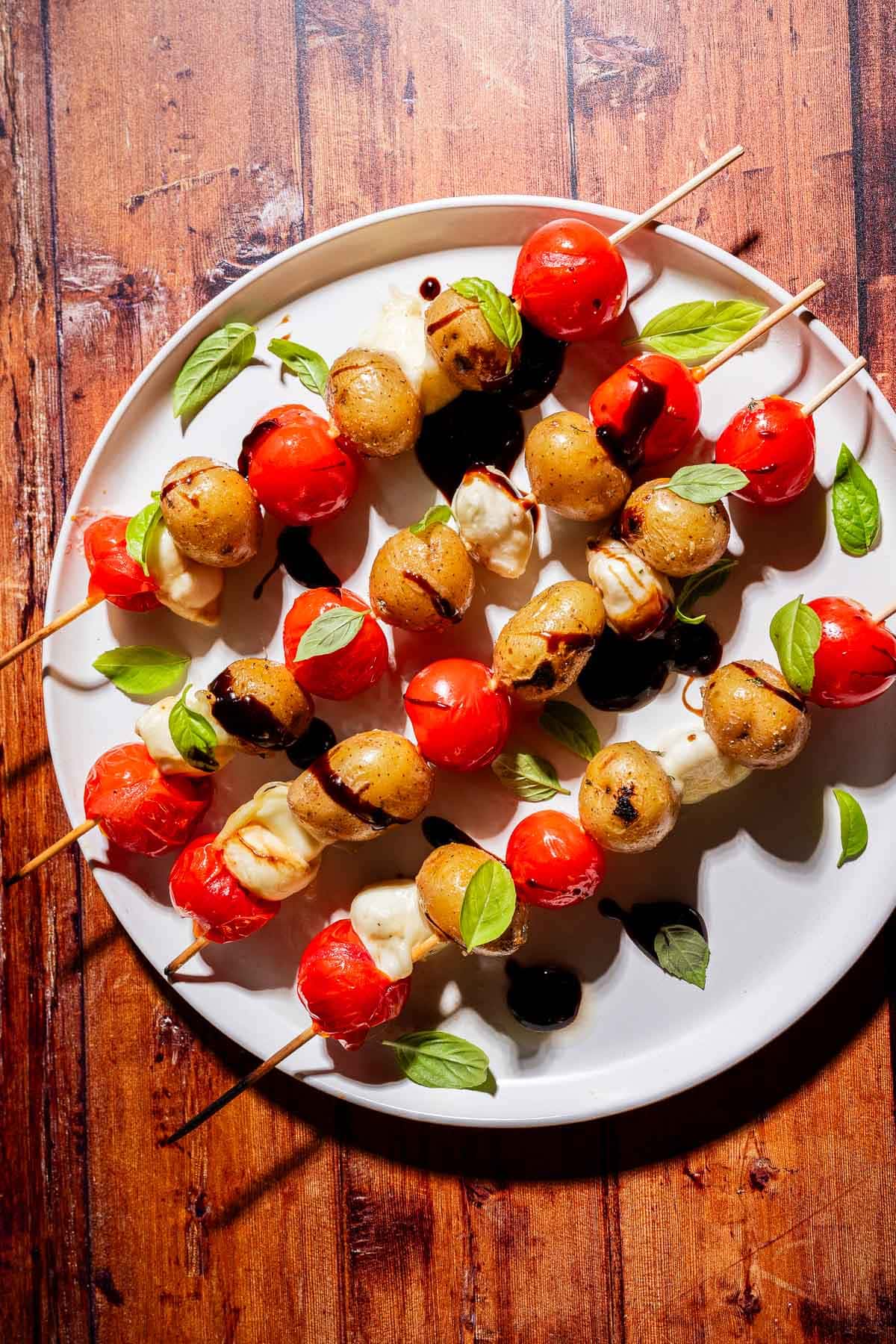 Skewers with cherry tomatoes, mozzarella balls, basil leaves, and a drizzle of balsamic glaze on a white plate atop a wooden surface.
