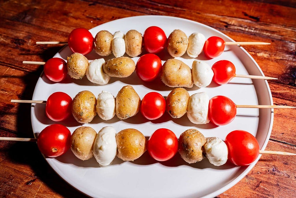 Four skewers on a white plate, each with cherry tomatoes, baby potatoes, and mozzarella balls, placed on a wooden surface.
