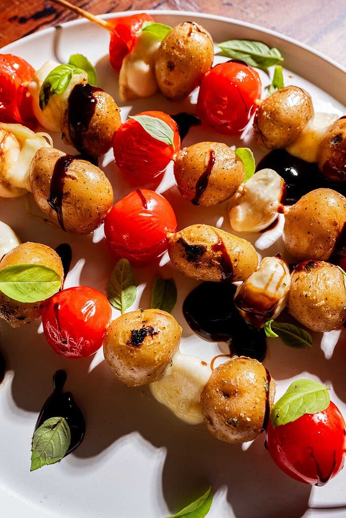 Skewers with grilled baby potatoes, cherry tomatoes, and mozzarella are garnished with fresh basil leaves and drizzled with balsamic glaze on a white plate.