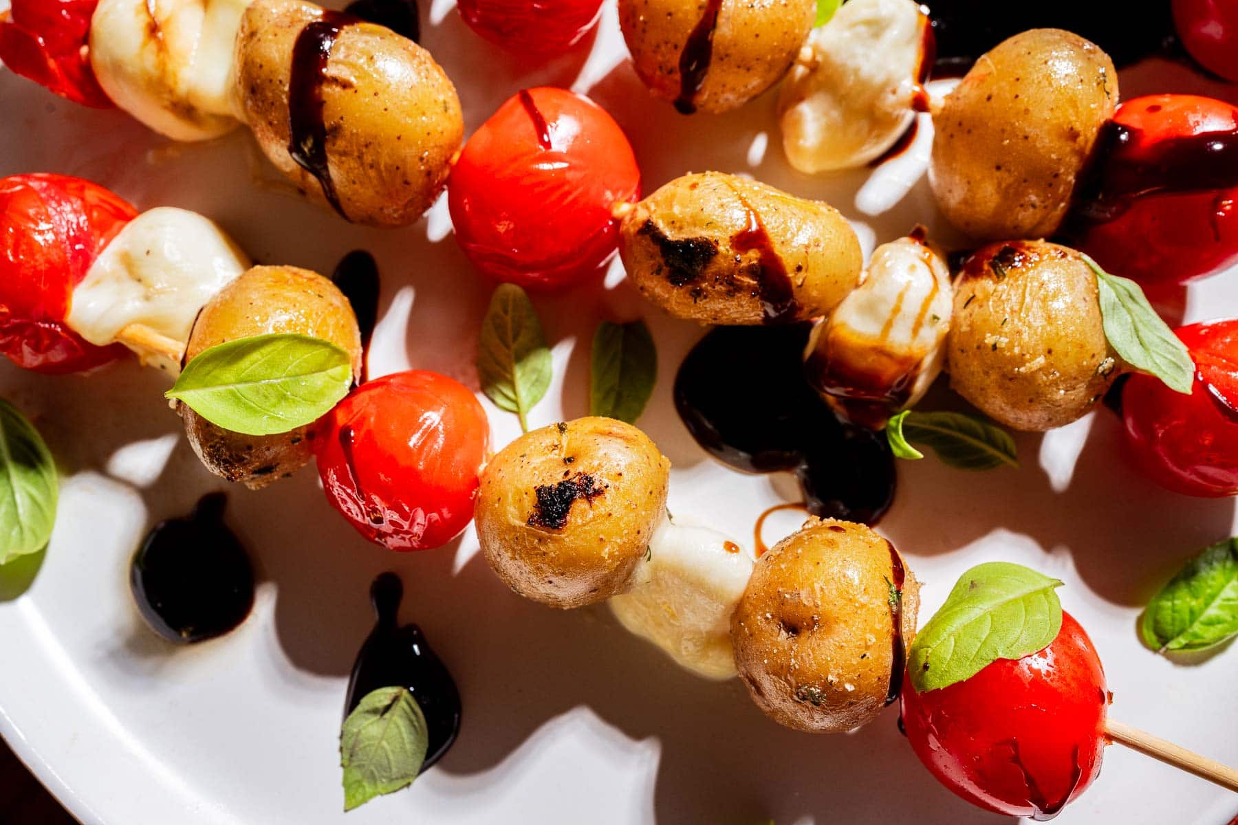 Skewers with roasted baby potatoes, cherry tomatoes, mozzarella, and basil leaves, drizzled with balsamic glaze on a white plate.