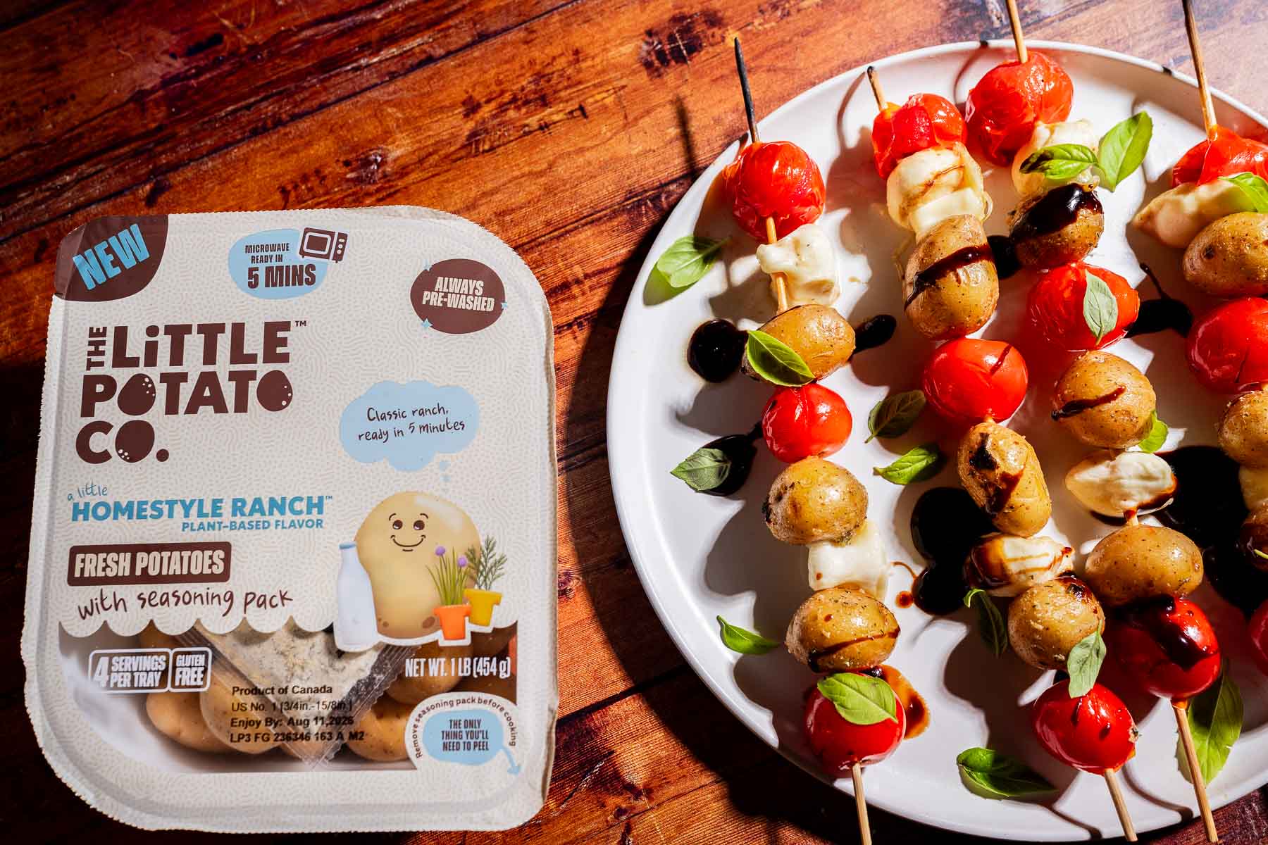 A package of The Little Potato Co. Homestyle Ranch potatoes sits next to skewers with cooked potatoes, cherry tomatoes, mozzarella balls, basil, and balsamic drizzle on a white plate.