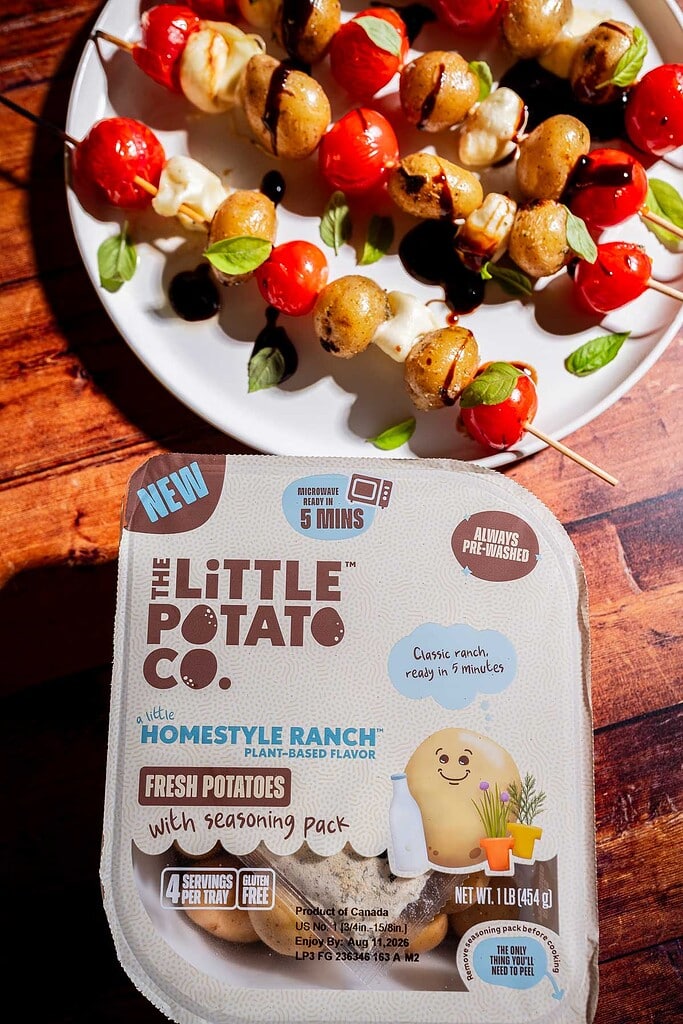 A package of The Little Potato Co. Homestyle Ranch potatoes sits on a wooden table next to a plate of skewered potatoes, cherry tomatoes, and mozzarella balls.
