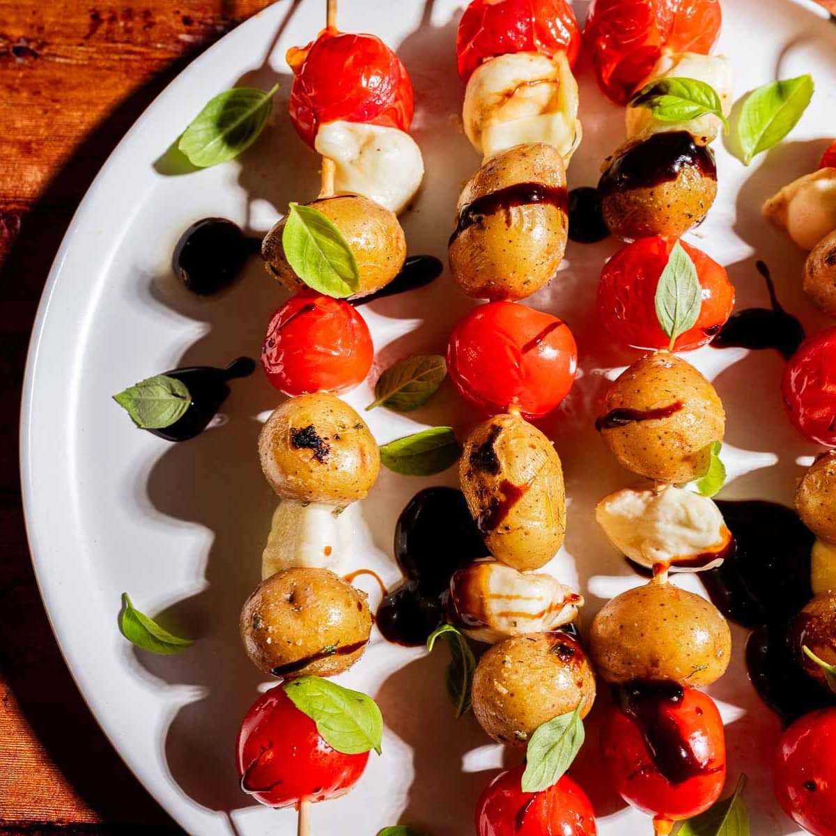 Skewers with roasted baby potatoes, cherry tomatoes, mozzarella, fresh basil, and balsamic glaze arranged on a white plate.