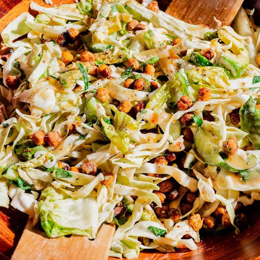 A hot honey salad of cabbage, sliced cucumbers, roasted chickpeas, and herbs is tossed in a wooden bowl with wooden salad servers.