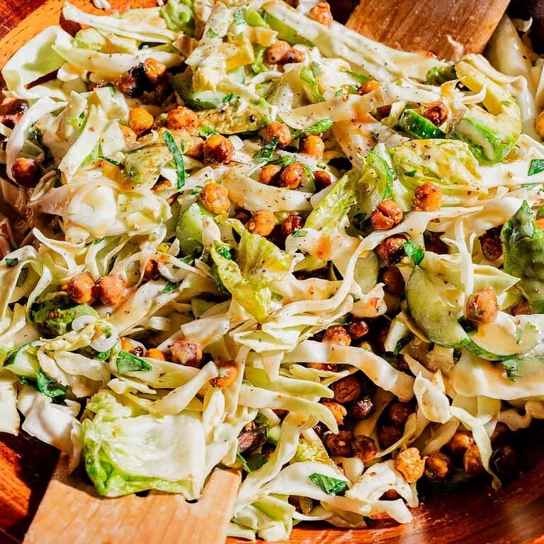 A hot honey salad of cabbage, sliced cucumbers, roasted chickpeas, and herbs is tossed in a wooden bowl with wooden salad servers.