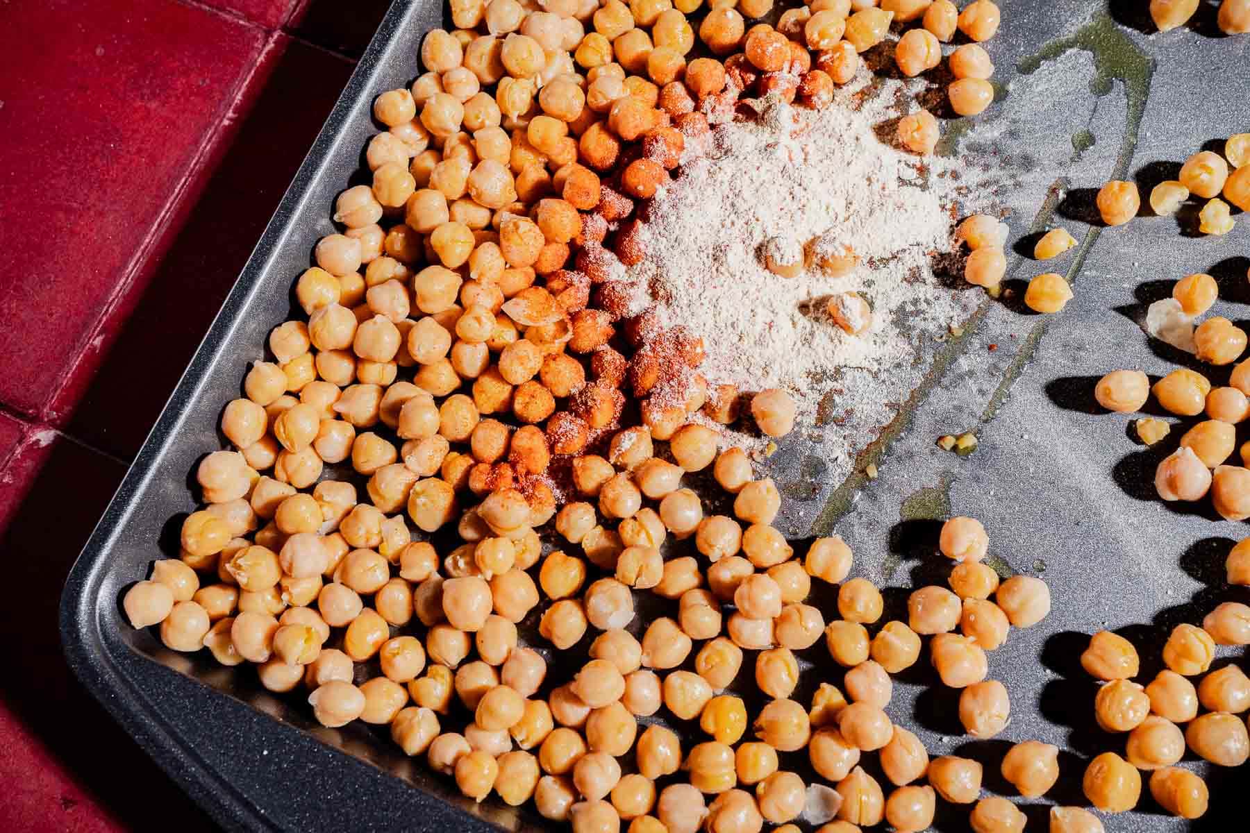 Chickpeas spread on a baking tray with oil, paprika, and flour, ready for roasting on a red tiled countertop—perfect for tossing into a hot honey salad.