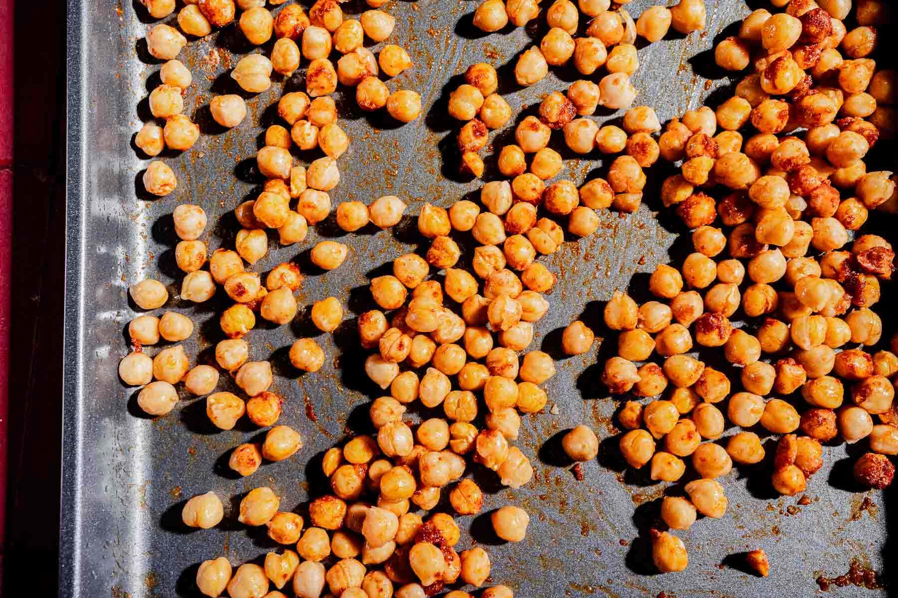 Roasted chickpeas scattered on a metal baking sheet, some with visible seasoning—perfect for tossing into a hot honey salad.