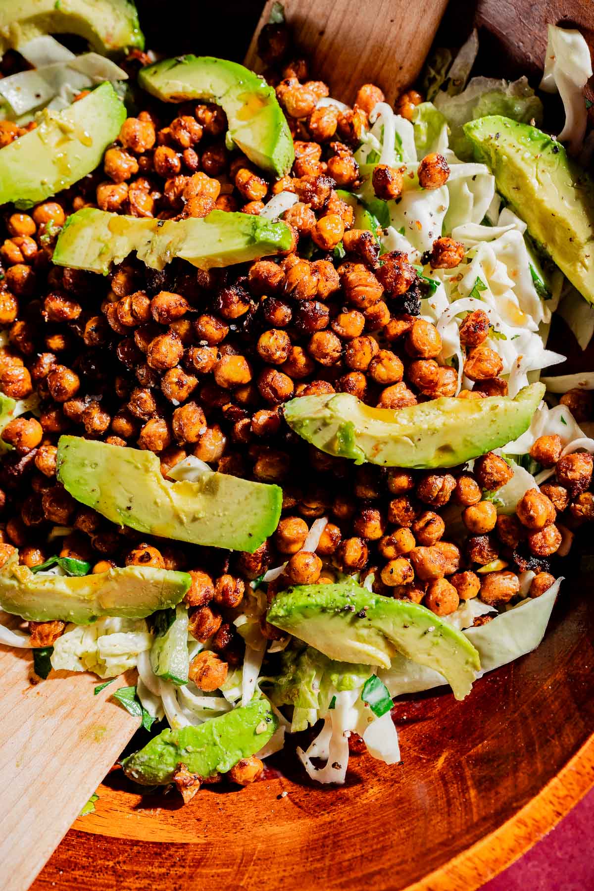 A wooden bowl filled with a hot honey salad of crispy roasted chickpeas, sliced avocado, chopped greens, and cabbage, with wooden serving utensils.