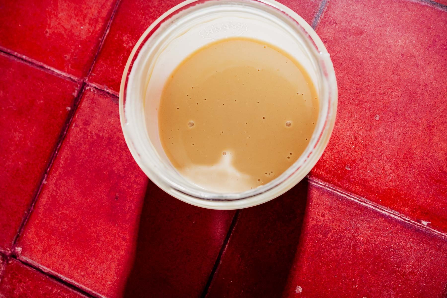 A clear glass bowl containing light brown hot honey salad sauce sits on a red tiled surface.
