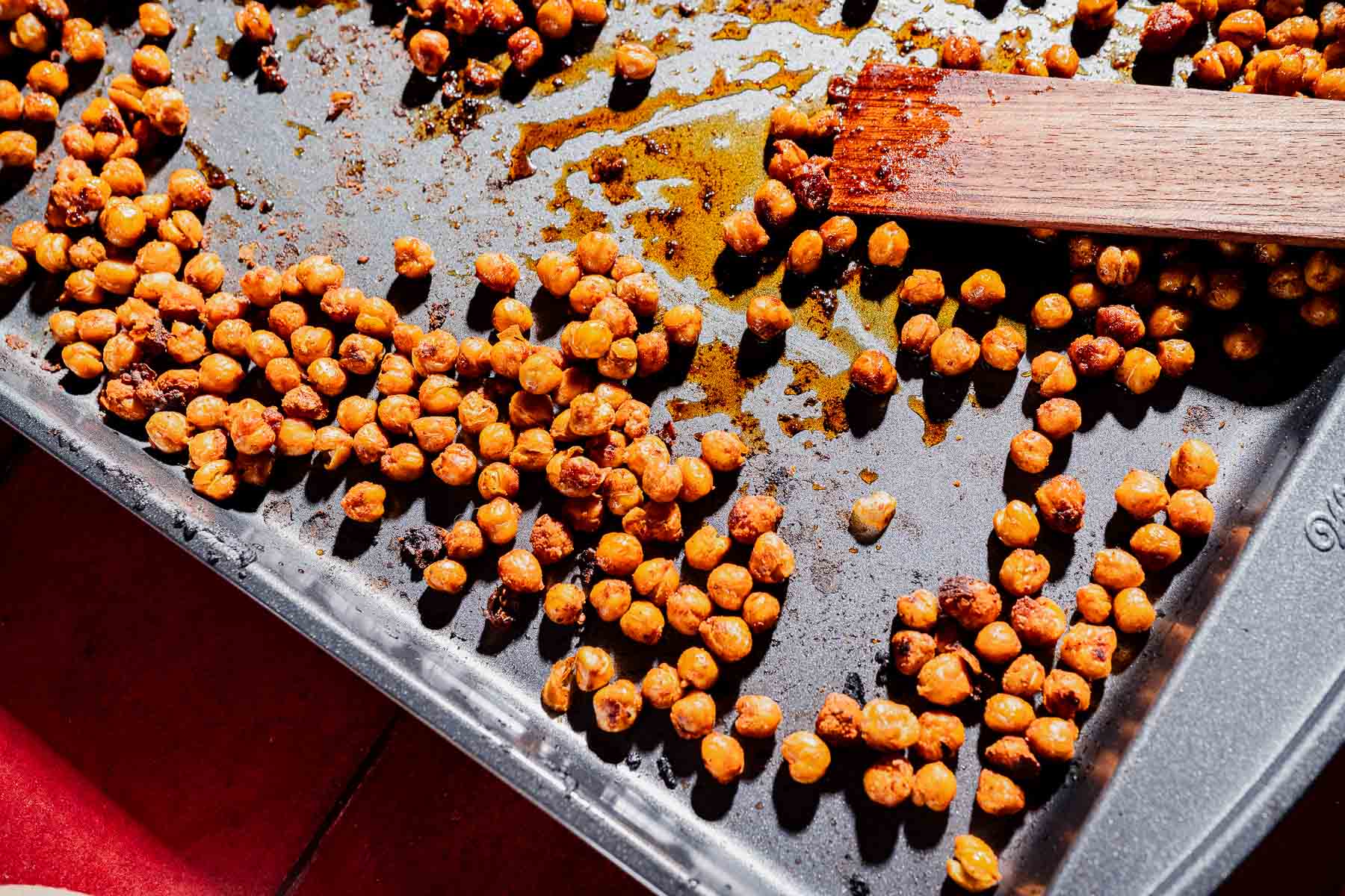 Seasoned roasted chickpeas scattered on a baking sheet, perfect for adding crunch and flavor to your next hot honey salad, with a wooden spatula resting on the edge.