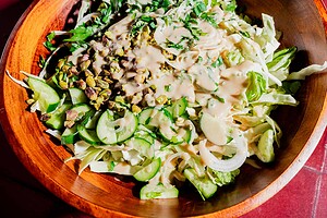 A wooden bowl filled with chopped lettuce, cabbage, cucumber, onions, pistachios, fresh herbs, and a creamy hot honey salad dressing.