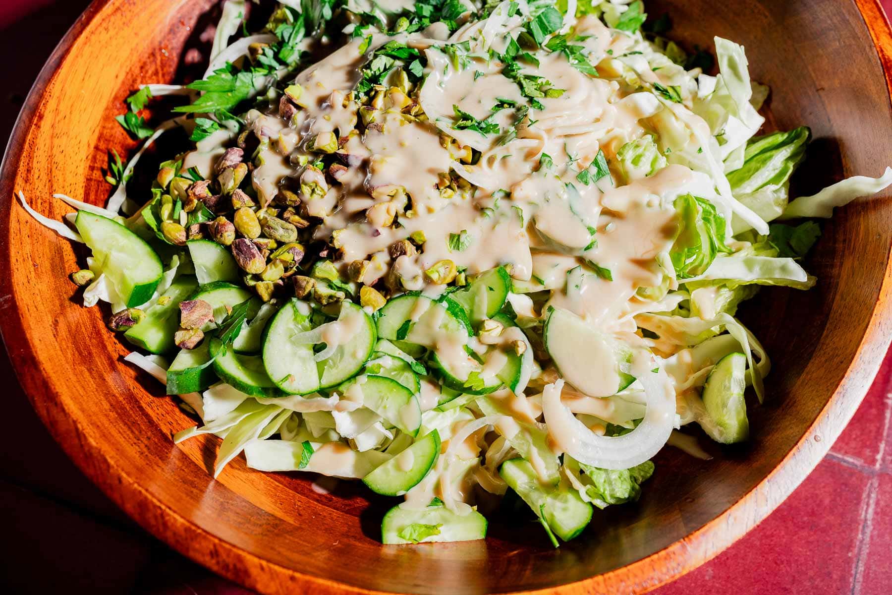 A wooden bowl filled with chopped lettuce, cabbage, cucumber, onions, pistachios, fresh herbs, and a creamy hot honey salad dressing.