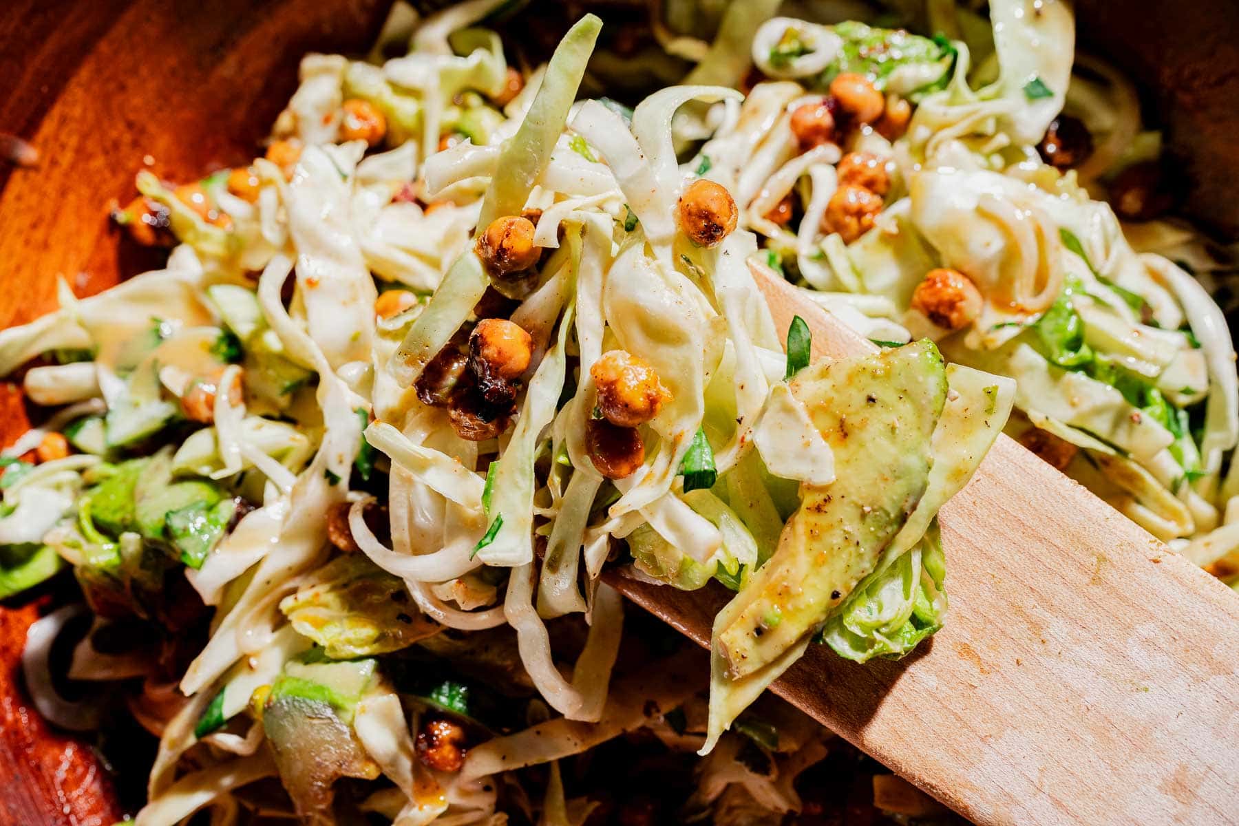 Shredded cabbage salad with avocado slices and roasted chickpeas, drizzled with hot honey, served in a wooden bowl with a wooden spatula.