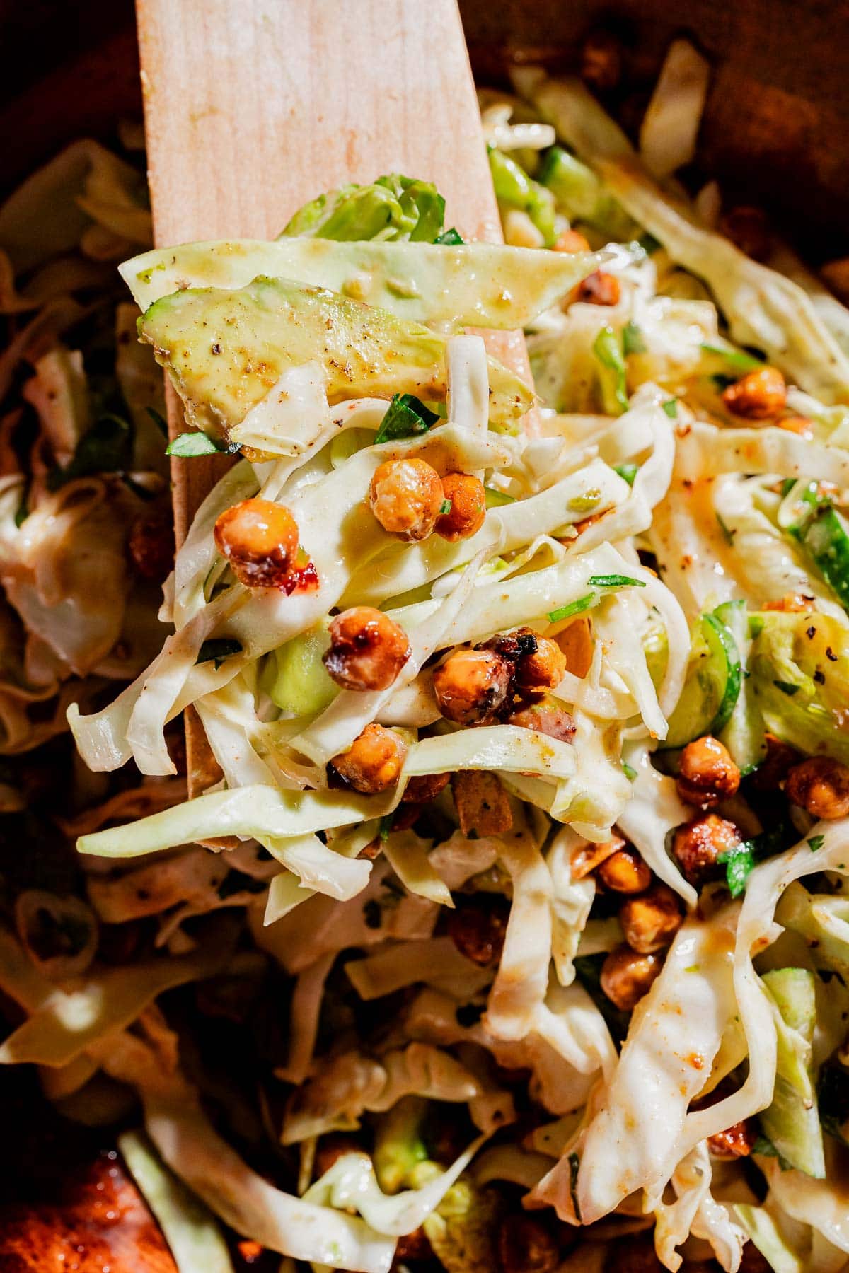 Close-up of a hot honey salad featuring cabbage, roasted chickpeas, avocado slices, and a creamy dressing being mixed with a wooden spatula.