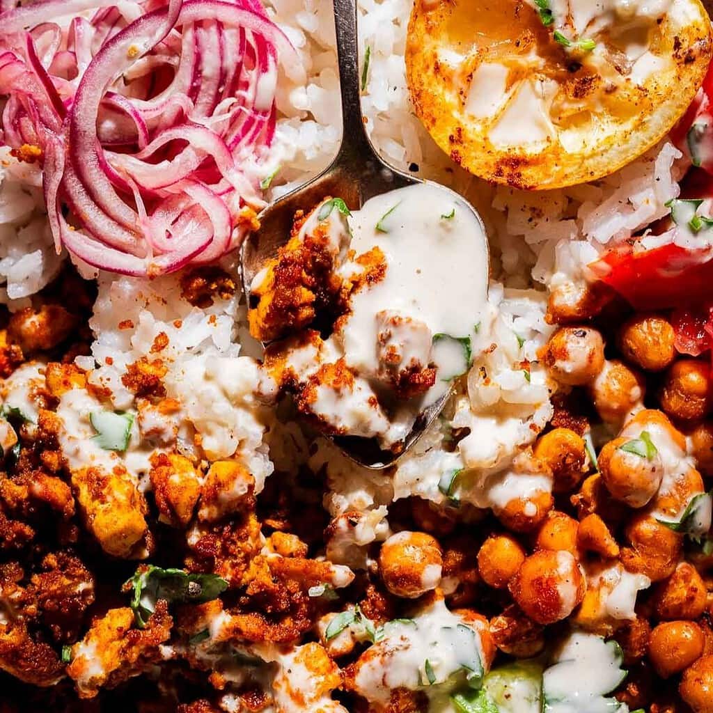 Close-up of a plate with white rice, roasted chickpeas, tofu shawarma, sliced red onions, a grilled lemon slice, fresh herbs, and creamy sauce, with a spoon scooping some of the food.