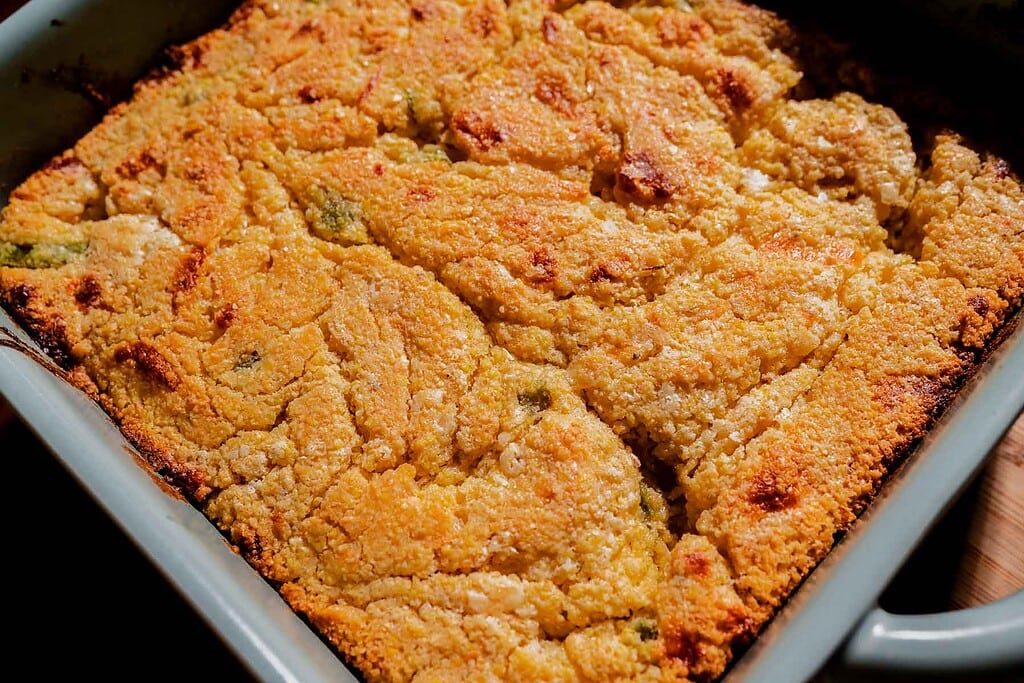 A close-up of a golden-brown cornbread with a slightly cracked surface, baked in a square metal pan.