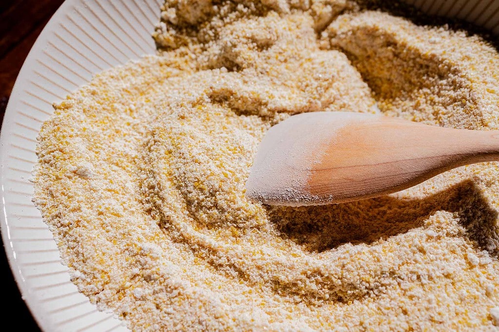 A wooden spoon resting in a bowl filled with dry yellow cornmeal.