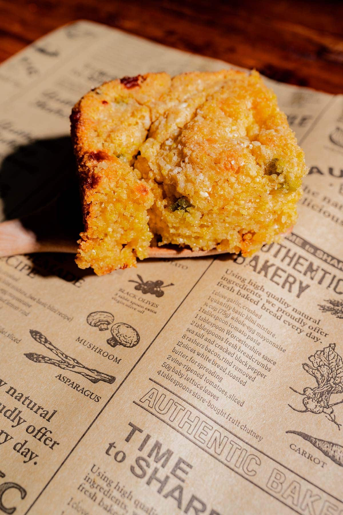 A close-up of a slice of cornbread with visible green chilies, resting on bakery-themed parchment paper.