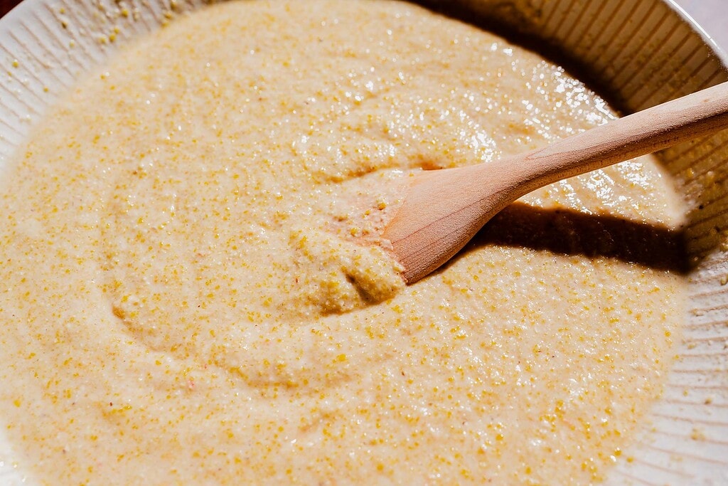 A wooden spoon stirs a bowl of thick, yellow cornmeal batter.