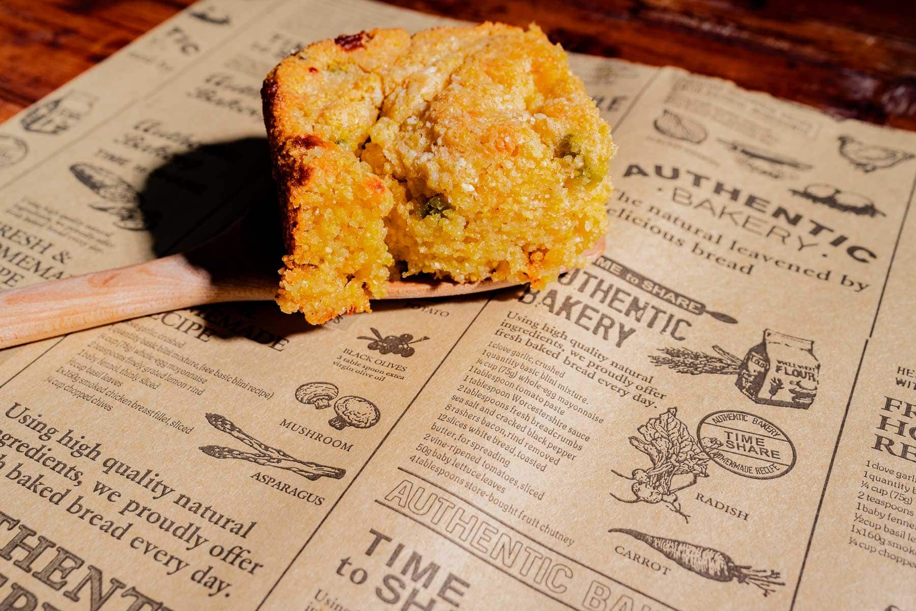 A slice of cornbread with visible green pieces rests on a wooden spatula over printed parchment paper with bakery-themed illustrations and text.