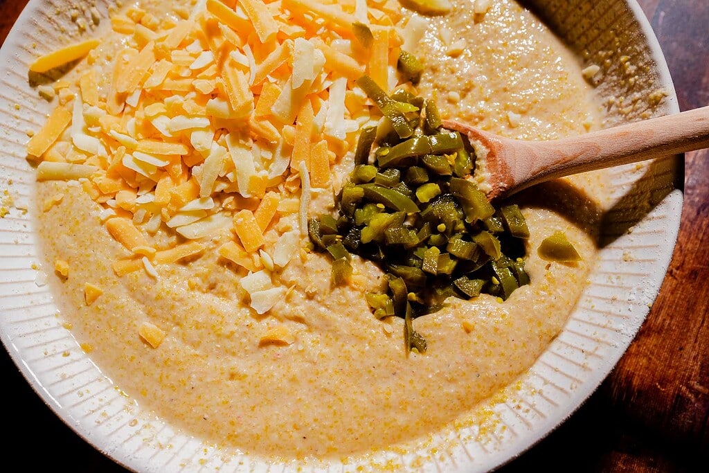 A bowl of creamy cornmeal mixture topped with shredded cheese and chopped green chilies, with a wooden spoon resting on the side.