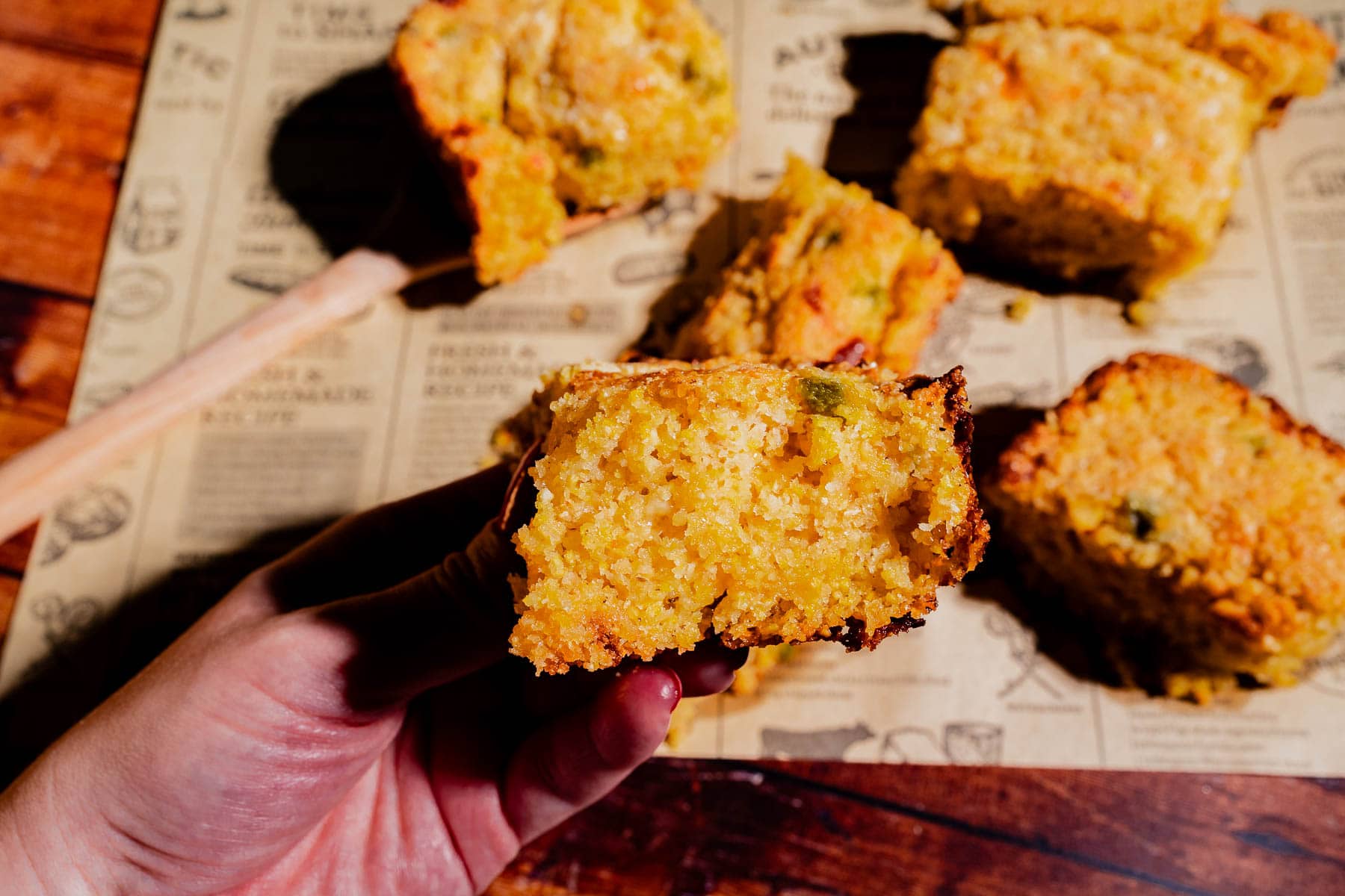A hand holds a piece of cornbread with a golden crust, with several more pieces of cornbread on printed paper in the background.
