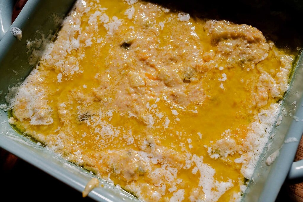 A close-up of an unbaked casserole in a pan, with a layer of melted butter and a mixture of batter and ingredients spread unevenly on top.